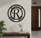 Satin black custom circular monogram metal plaque featuring the initial letter R, displayed on an indoor white stucco partition wall above a chunky rustic wooden bench table holding pampas grass.