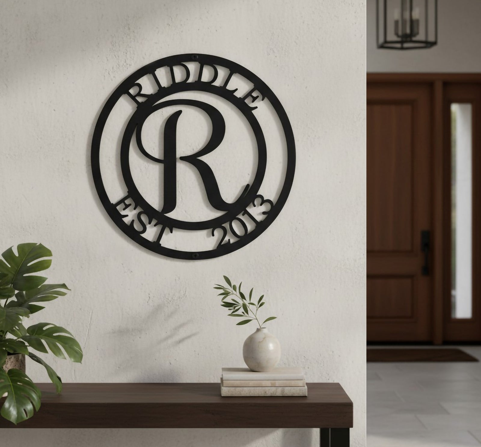 Satin black custom circular monogram metal plaque featuring the initial letter R, displayed on an indoor white stucco partition wall above a chunky rustic wooden bench table holding pampas grass.