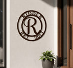 Rich espresso brown finish of the personalized round metal initial monogram sign featuring the surname Riddle, mounted on a white outdoor building wall next to a wooden doorframe and a dark planter pot.