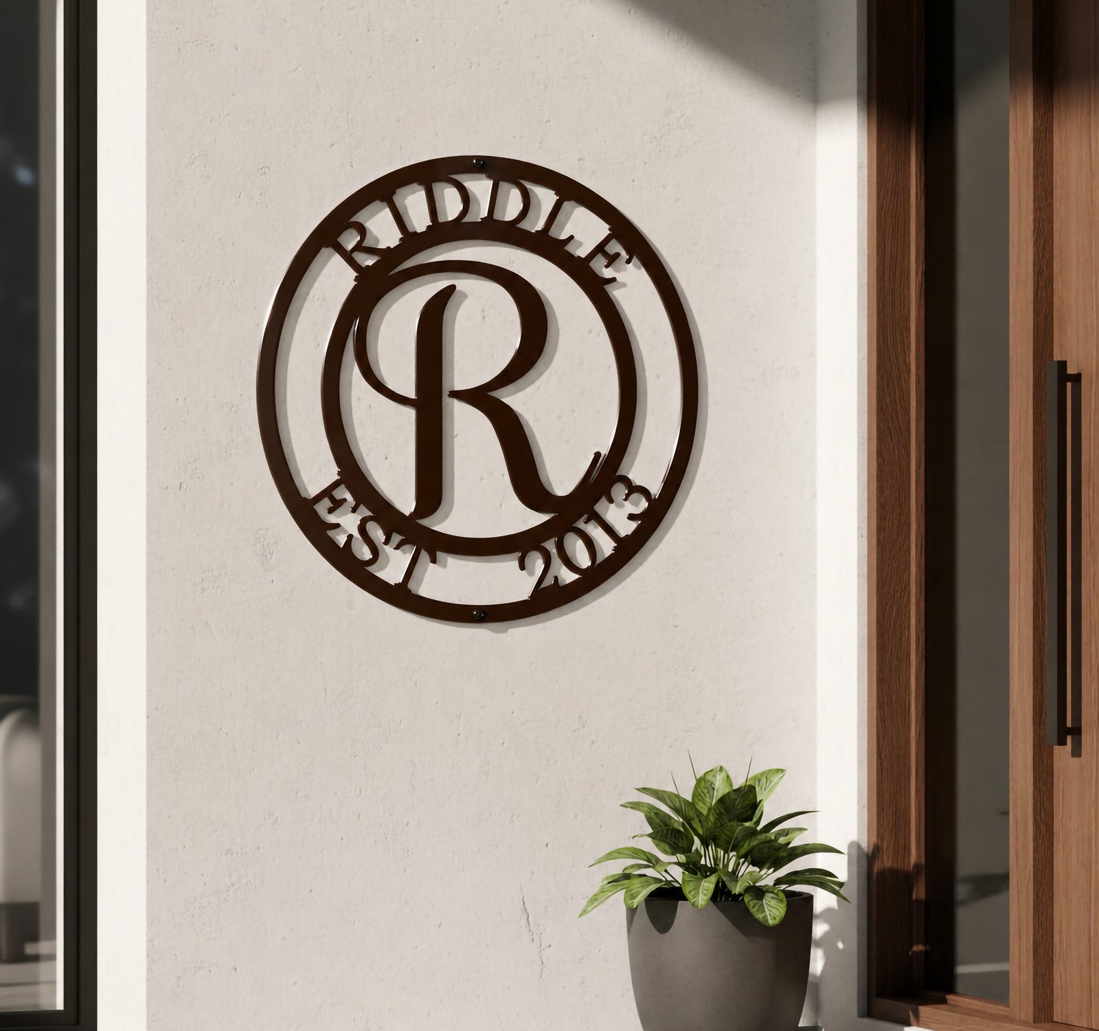 Rich espresso brown finish of the personalized round metal initial monogram sign featuring the surname Riddle, mounted on a white outdoor building wall next to a wooden doorframe and a dark planter pot.