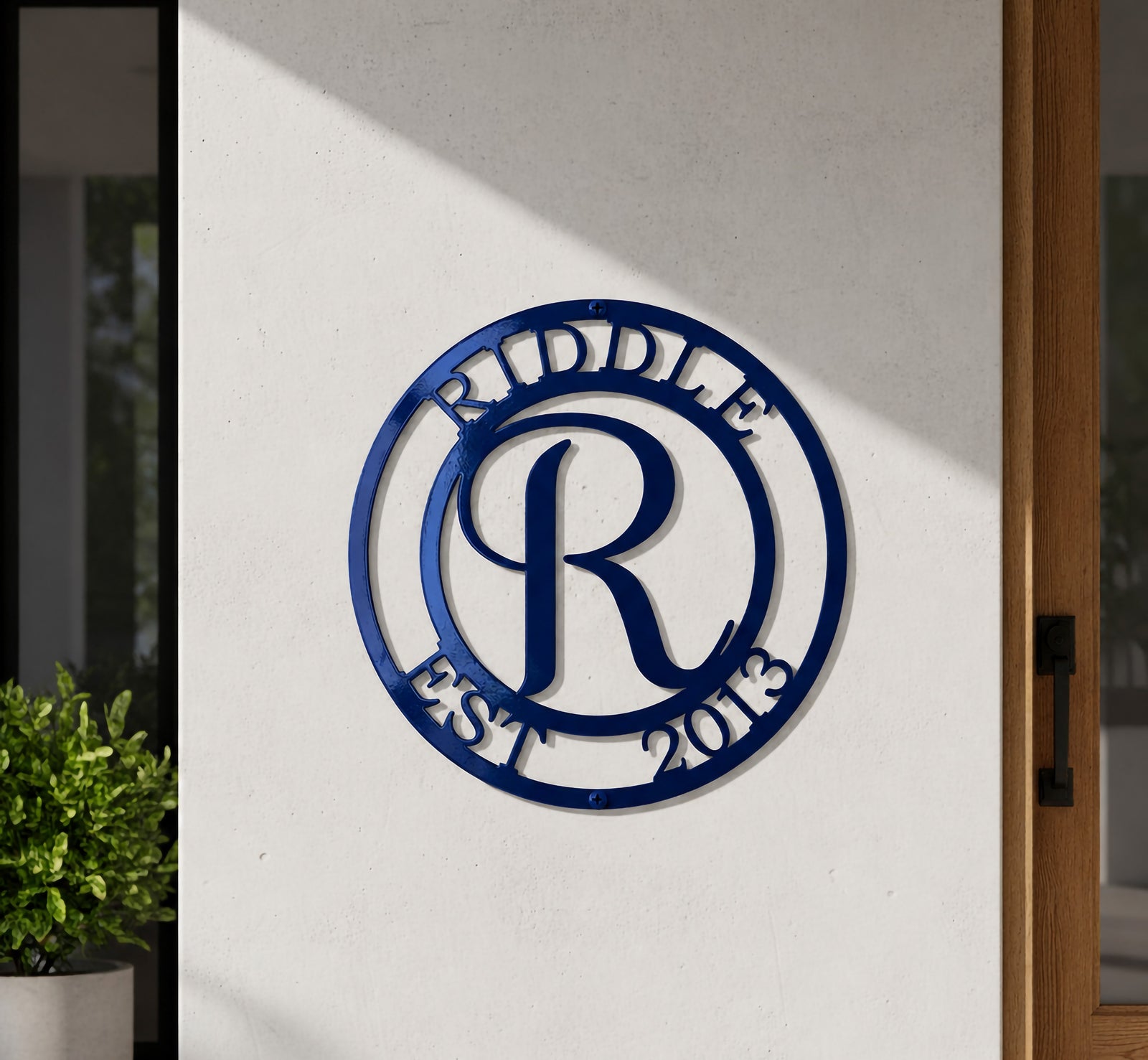 Glossy cobalt blue finish of the custom circular family name monogram sign with custom text Riddle Est 2013, mounted flat on a white exterior entry wall beside a warm wood-framed glass front door.