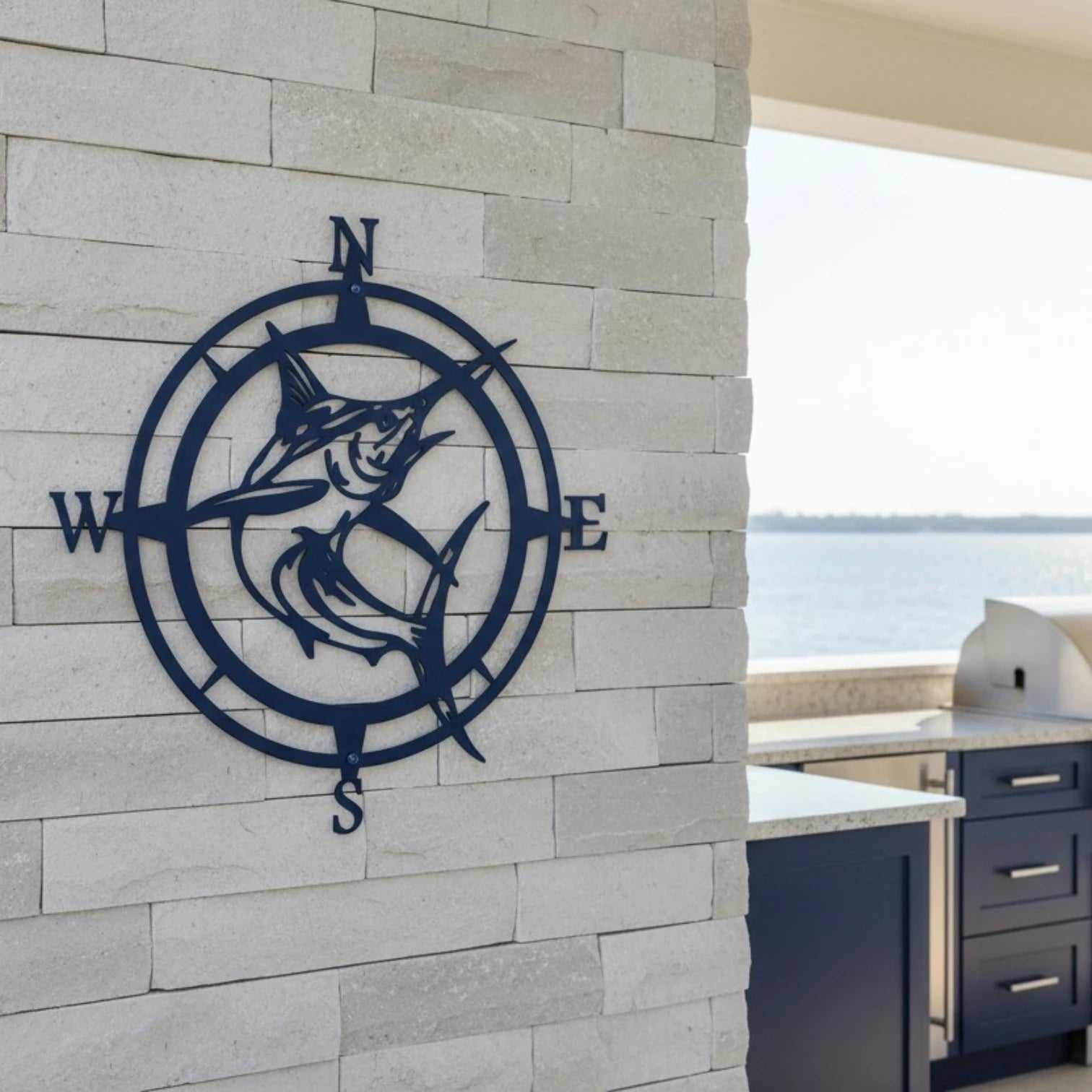Dark navy blue metal marlin compass rose sign mounted on a light gray stacked stone accent wall of a luxury outdoor kitchen patio.