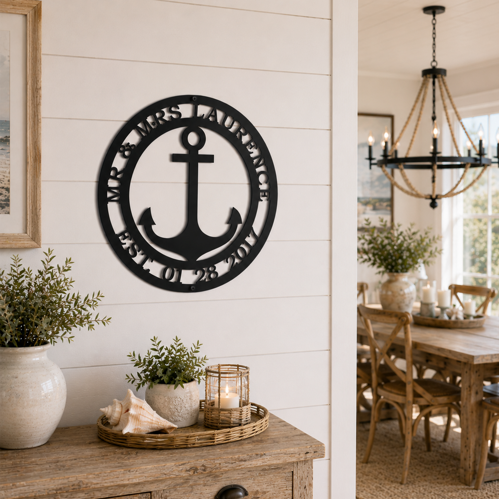 Custom black metal anchor family name sign displayed on an interior white entryway wall next to a coastal dining room setup with a rope chandelier.