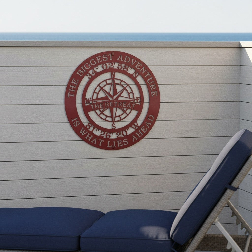 A crimson red powder-coated circular metal compass wall hanging is mounted outdoors on a gray-tinted horizontal wood slat wall. The sign displays custom latitude and longitude coordinates along with "THE RETREAT" in the center. Part of a blue and white patio lounge chair faces the ocean backdrop.