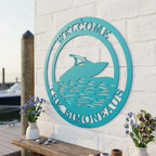 A teal round metal sign customized with "Welcome The Simoneaus" and a boat silhouette, fixed to a white textured brick wall on a marina dock.