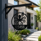 A circular matte black metal address sign featuring a laser-cut octopus motif above the numbers "123456". The sign hangs from short chains attached to a black metal wall bracket mounted on a dark grey stucco pillar, showcasing a concrete walkway and landscaped front yard in the background.