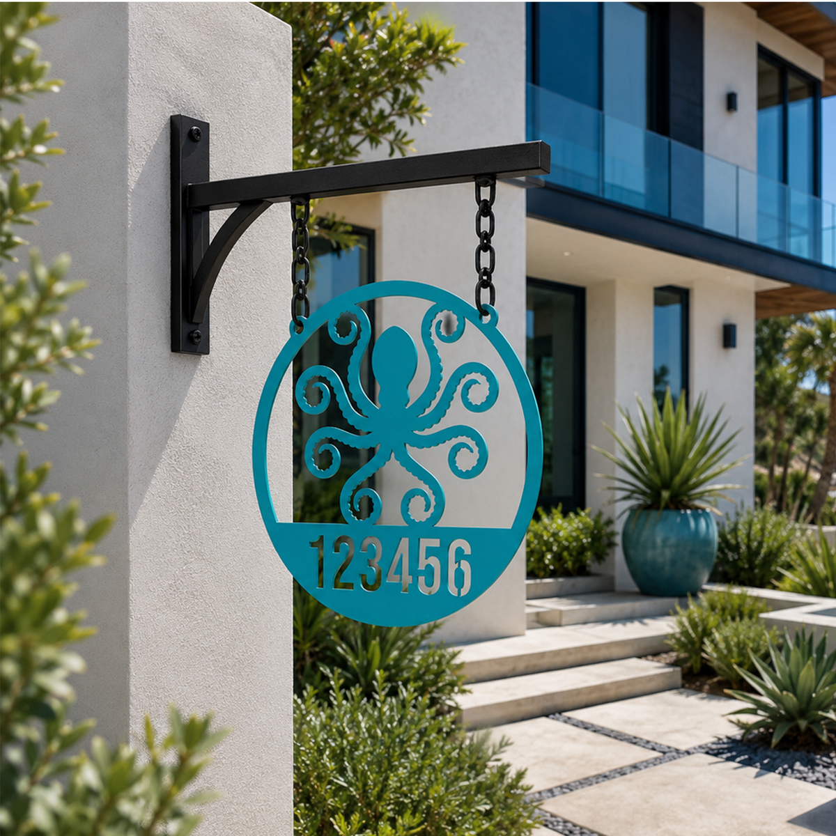 A circular custom metal house sign in a vibrant turquoise finish, highlighting a laser-cut octopus shape and the custom text "123456". The piece hangs from chains on a black metal post attached to a textured white stucco entry wall of a contemporary home.