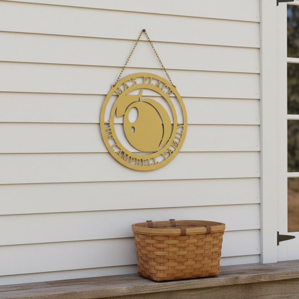 Gold-colored personalized round metal peach orchard sign hanging from a chain on a white shiplap exterior wall above a woven wooden harvest basket.