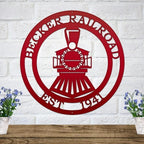 Red metal steam train sign with custom name and date on white brick above a wooden shelf with blue potted flowers.