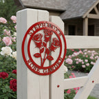 Deep candy apple red finish of the custom circular metal rose sign mounted directly on a white wooden gate post at the entrance of a garden path with a rustic log home behind it.