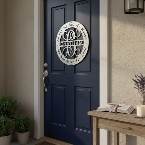 A clean matte white circular metal monogram sign featuring the name "CHATHAM" and a scripture verse is hung directly on a navy blue front door. The scene features a welcoming porch with potted green shrubs and a small wooden side table holding an open Bible and a lit candle.