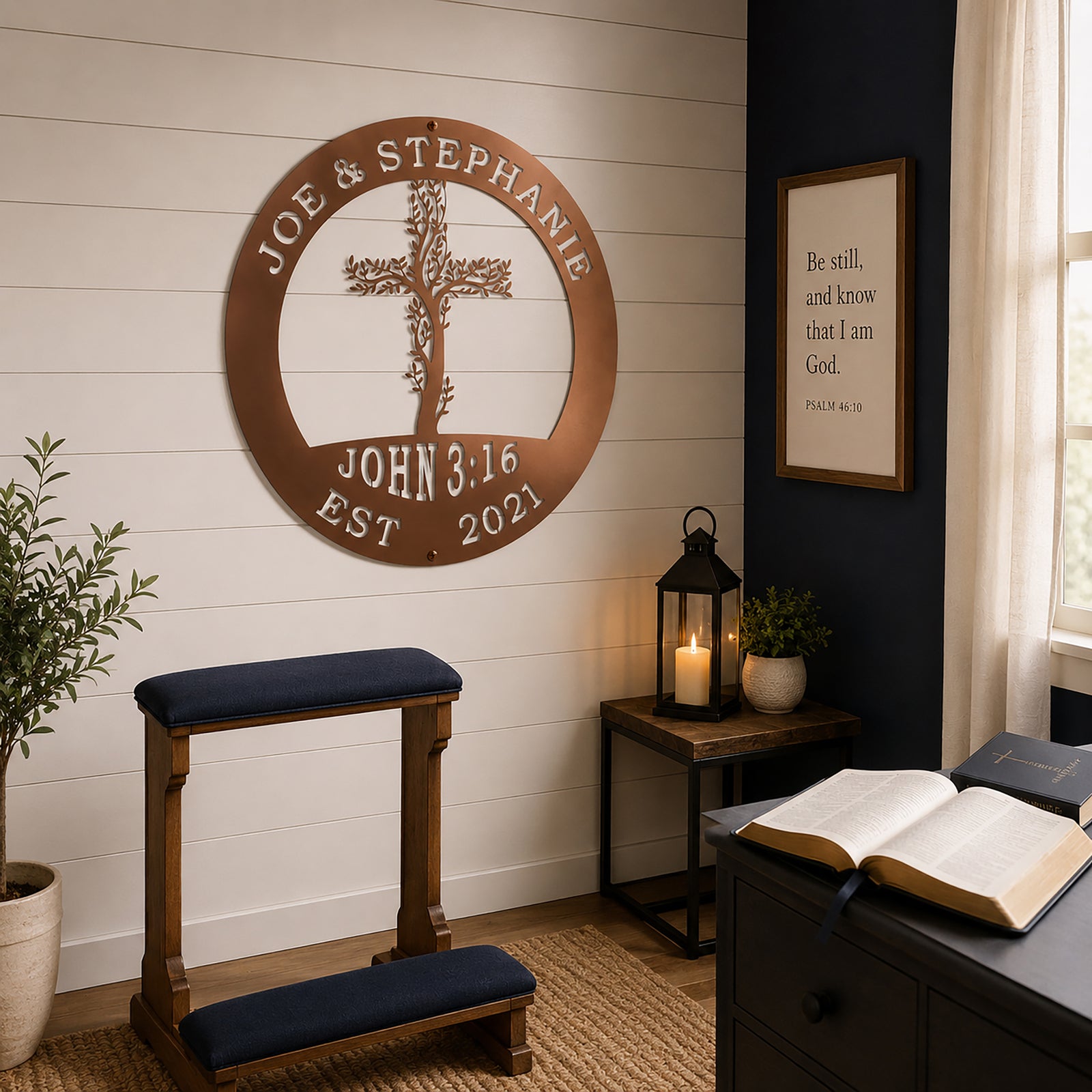 A metallic rose gold circular metal sign displaying the custom text "JOE & STEPHANIE" and "JOHN 3:16" is hung in a prayer room setting. The sign is mounted on white wood paneling over a wooden kneeling bench, a side table with a lit candle lantern, and an open Bible resting on a nearby desk.