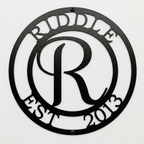 Flat view of a textured black circular personalized metal sign featuring a large script letter R monogram, cut out with the family name Riddle and established date 2013 over a pure white background.