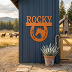 Orange Rocky horse horseshoe metal sign with horse silhouette displayed on blue barn siding near a ranch gate and pasture
