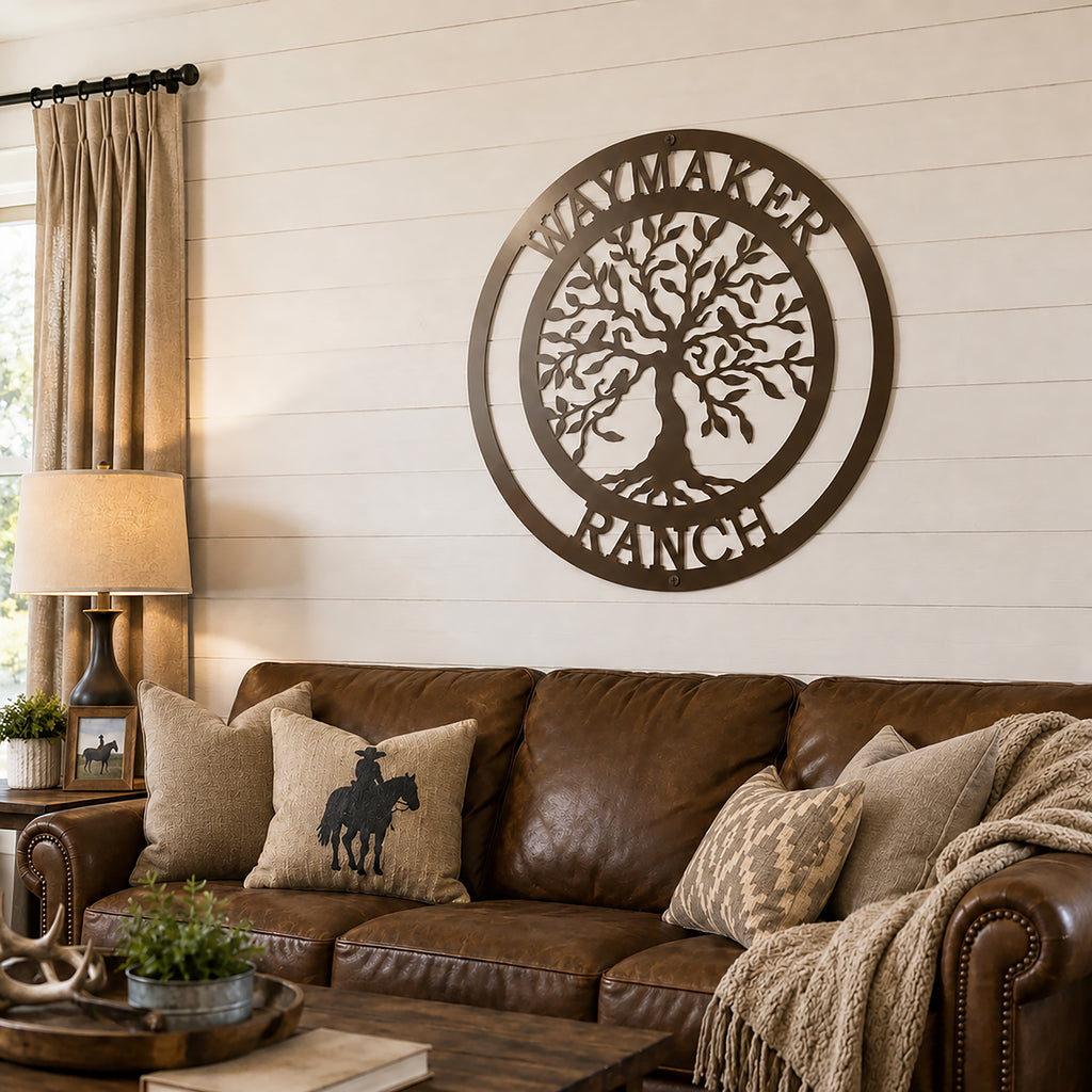 A rich espresso brown circular metal sign showcasing a Tree of Life and custom ranch name is hung on a white shiplap living room wall. The Western-themed room features a brown leather sofa styled with cowboy-themed accent pillows, tan drapes, and a dark wood coffee table.