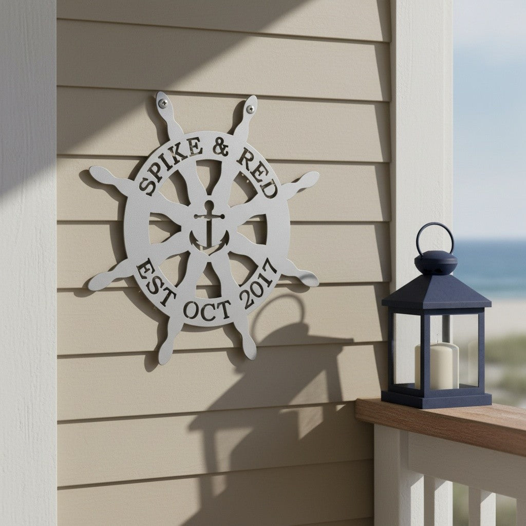 Textured white personalized ship steering wheel metal monogram sign featuring an anchor design and custom text, mounted on a tan shiplap wall next to a blue candle lantern.