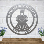 Silver metal train sign featuring custom text on a white brick wall above a rustic shelf with blue flowers in pots.