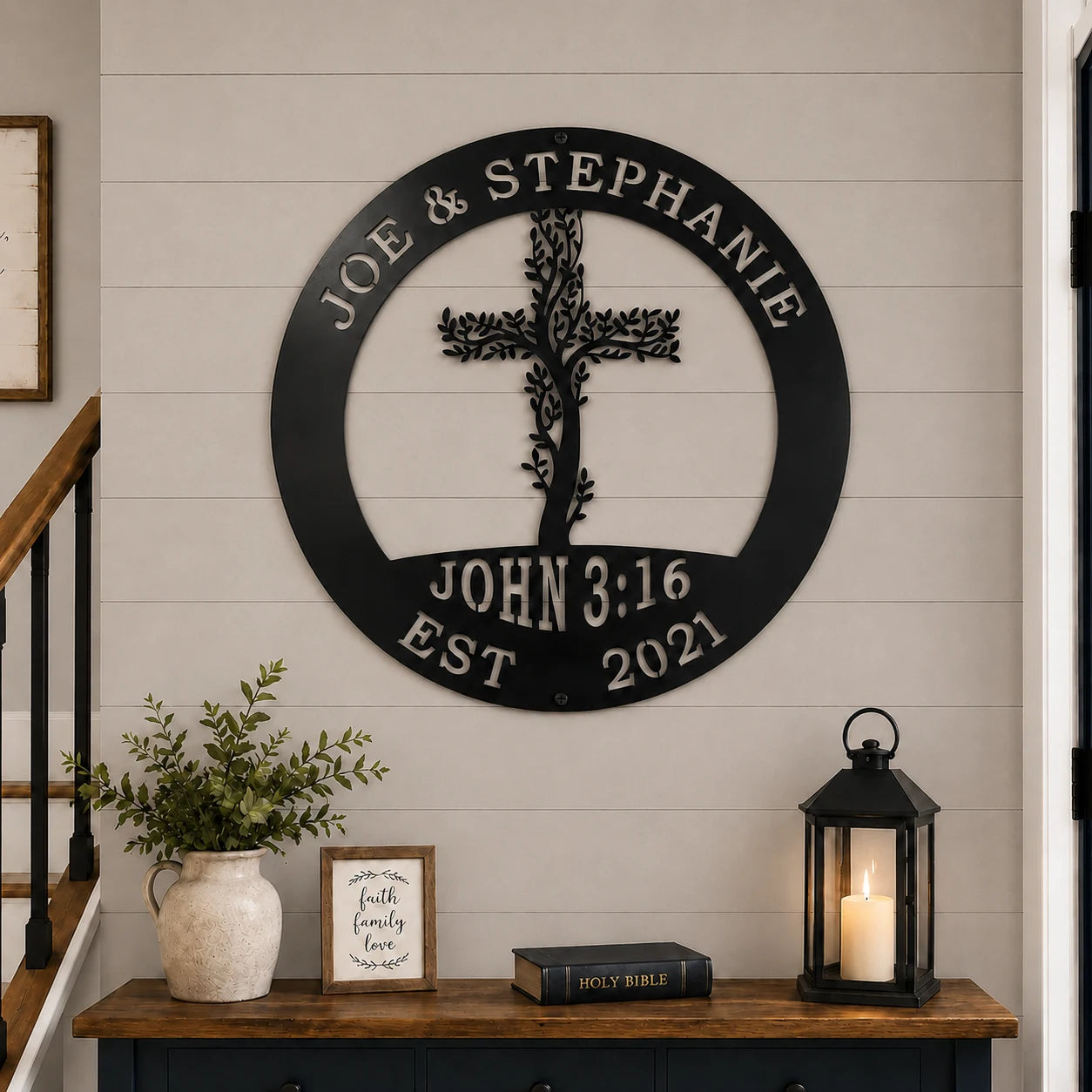 Personalized black metal wall art featuring a cross and the text 'JOE & STEPHANIE' and 'JOHN 3:16 EST 2021', displayed on a white shiplap wall above a wooden console table with decorative items.