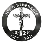 A circular metal sign finished in a glossy black color is displayed over a solid white background. The top rim is cut out with the names "JOE & STEPHANIE," the bottom features "JOHN 3:16 EST 2021," and the center showcases an intricate cross woven with climbing tree branches and leaves.