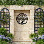 Black round laser-cut metal tree of life sign customized with McCallum displayed inside a stone wall alcove flanked by blue wooden trellises and potted purple hydrangea flowers.