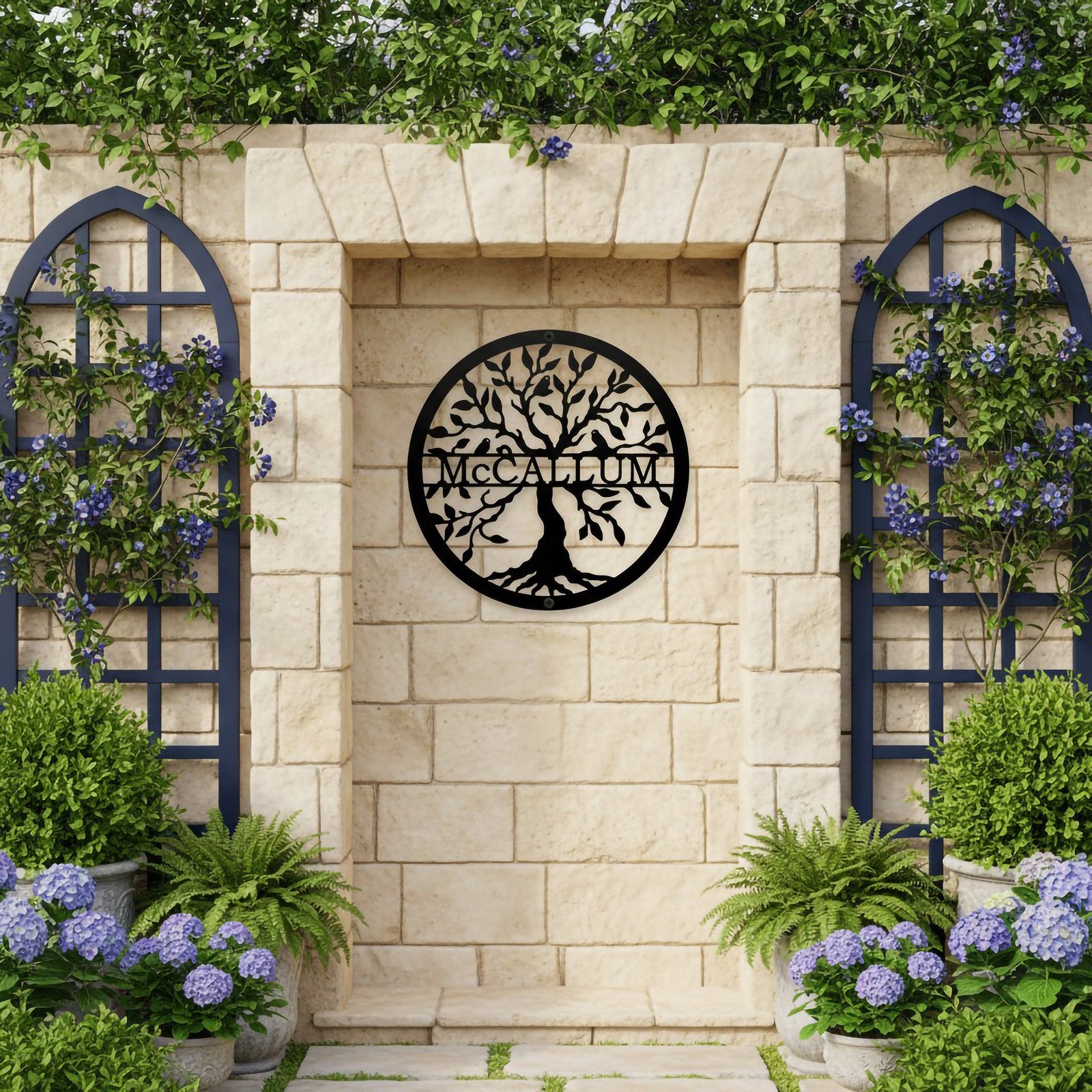 Black round laser-cut metal tree of life sign customized with McCallum displayed inside a stone wall alcove flanked by blue wooden trellises and potted purple hydrangea flowers.