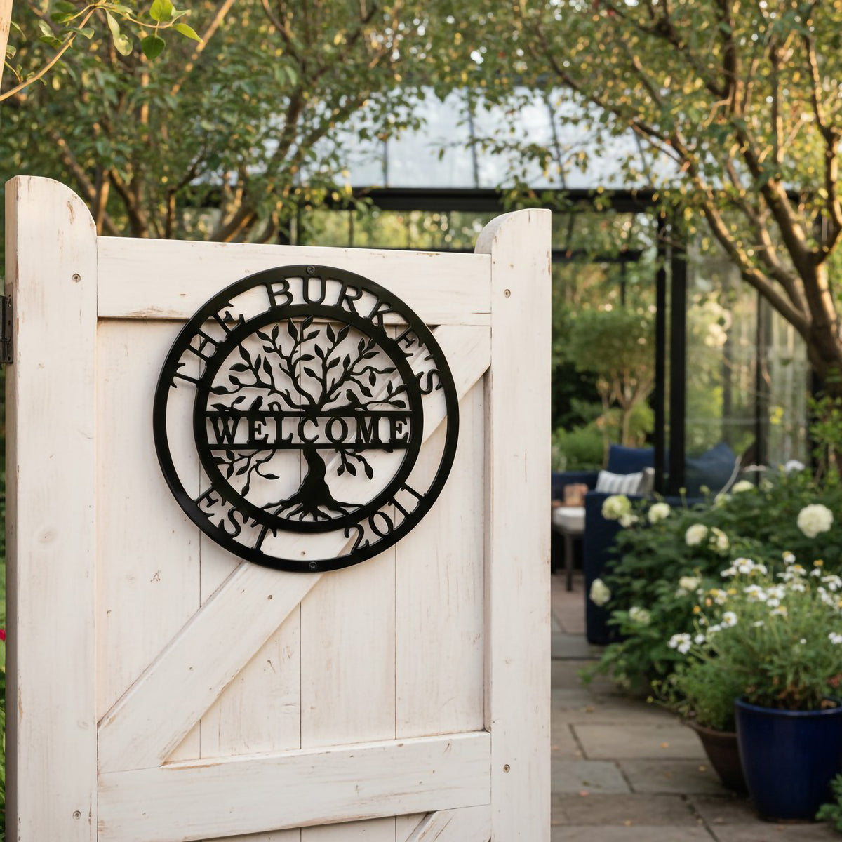 A black round metal family sign reading "The Burkes Welcome Est. 2011" featuring a detailed tree motif, fixed to an open white wooden garden gate leading into a lush patio.
