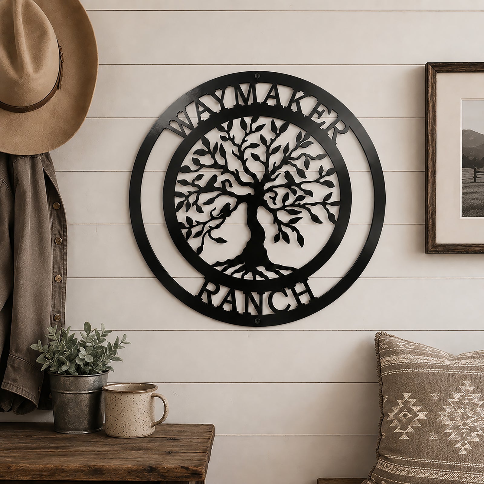Classic black personalized "Waymaker Ranch" tree of life metal sign displayed on an indoor white shiplap wall next to a hanging cowboy hat and a small rustic table.