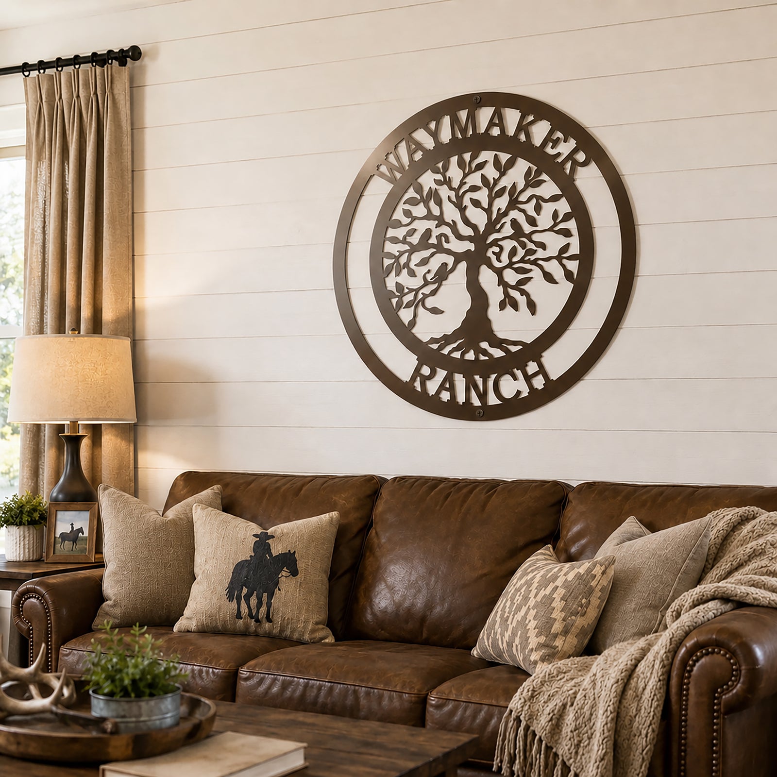 Espresso brown personalized metal tree of life wall art reading "Waymaker Ranch", displayed on white shiplap above a brown leather couch in a rustic living room
