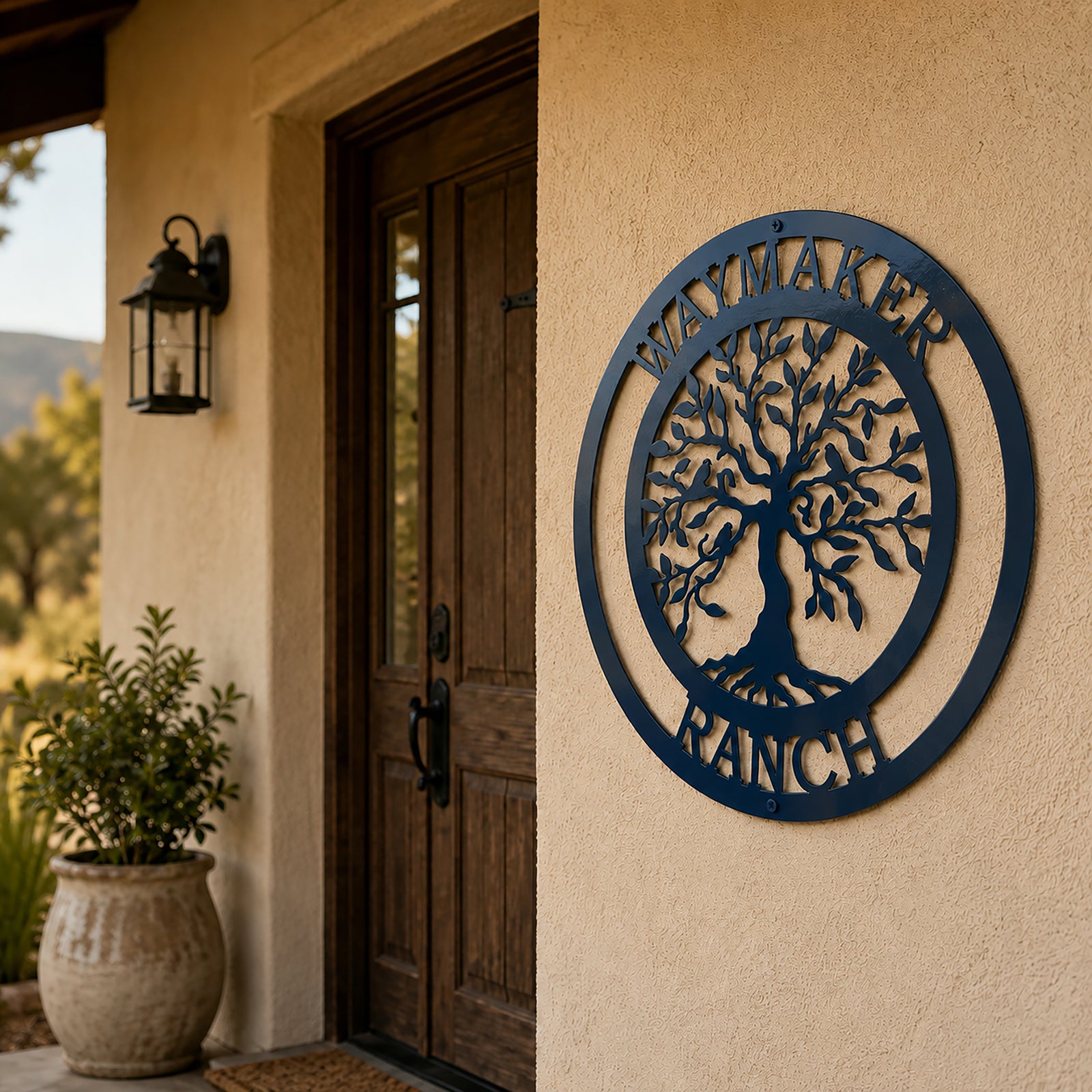 Navy blue custom "Waymaker Ranch" metal tree of life sign hung on an exterior stucco wall next to a wooden front entryway door and a potted plant.