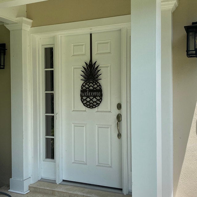 Black pineapple welcome metal door hanger displayed on white front door for inviting porch entry decor