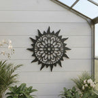 A dark grey metal sun medallion featuring a detailed inner floral cutout, mounted on a white shiplap wall inside a bright greenhouse surrounded by potted tropical plants.