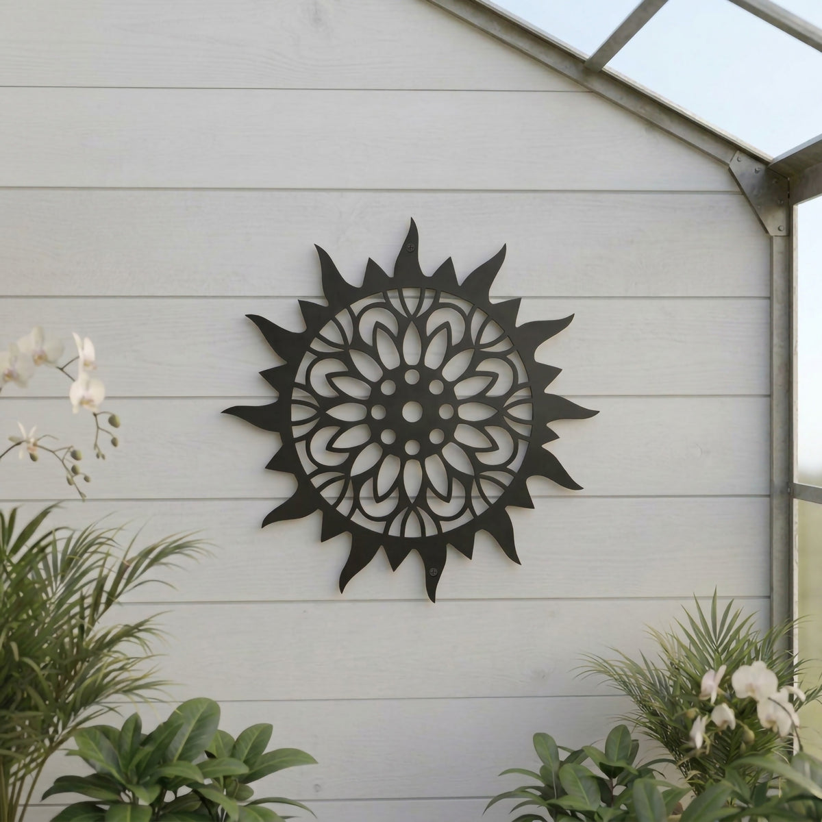 A dark grey metal sun medallion featuring a detailed inner floral cutout, mounted on a white shiplap wall inside a bright greenhouse surrounded by potted tropical plants.