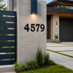 A bold black metal house numbers displaying "4579" are installed on a light-colored masonry pillar under a glowing accent light. A contemporary home exterior with a dark garage door, stone tile details, and a geometric concrete driveway is visible in the background.