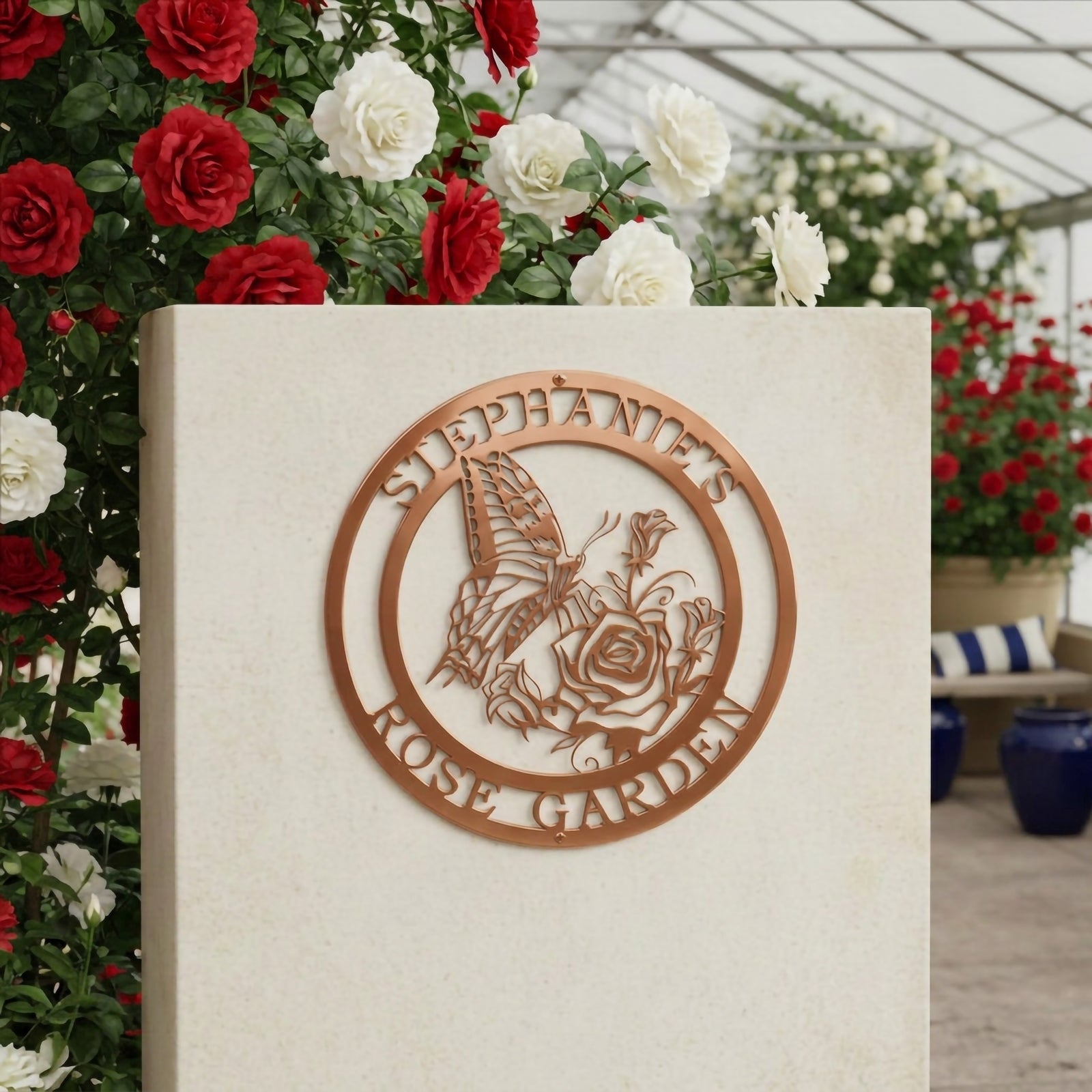 Metallic rose gold finish of the custom circular butterfly rose sign mounted on a smooth indoor presentation column wall surrounded by red and white rose bushes inside a bright garden sunroom.