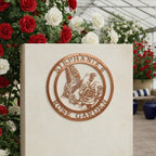 A copper-colored metallic circular sign reading "Stephanie's Rose Garden," displayed on a smooth light stone pedestal inside a greenhouse surrounded by vibrant red and white roses.