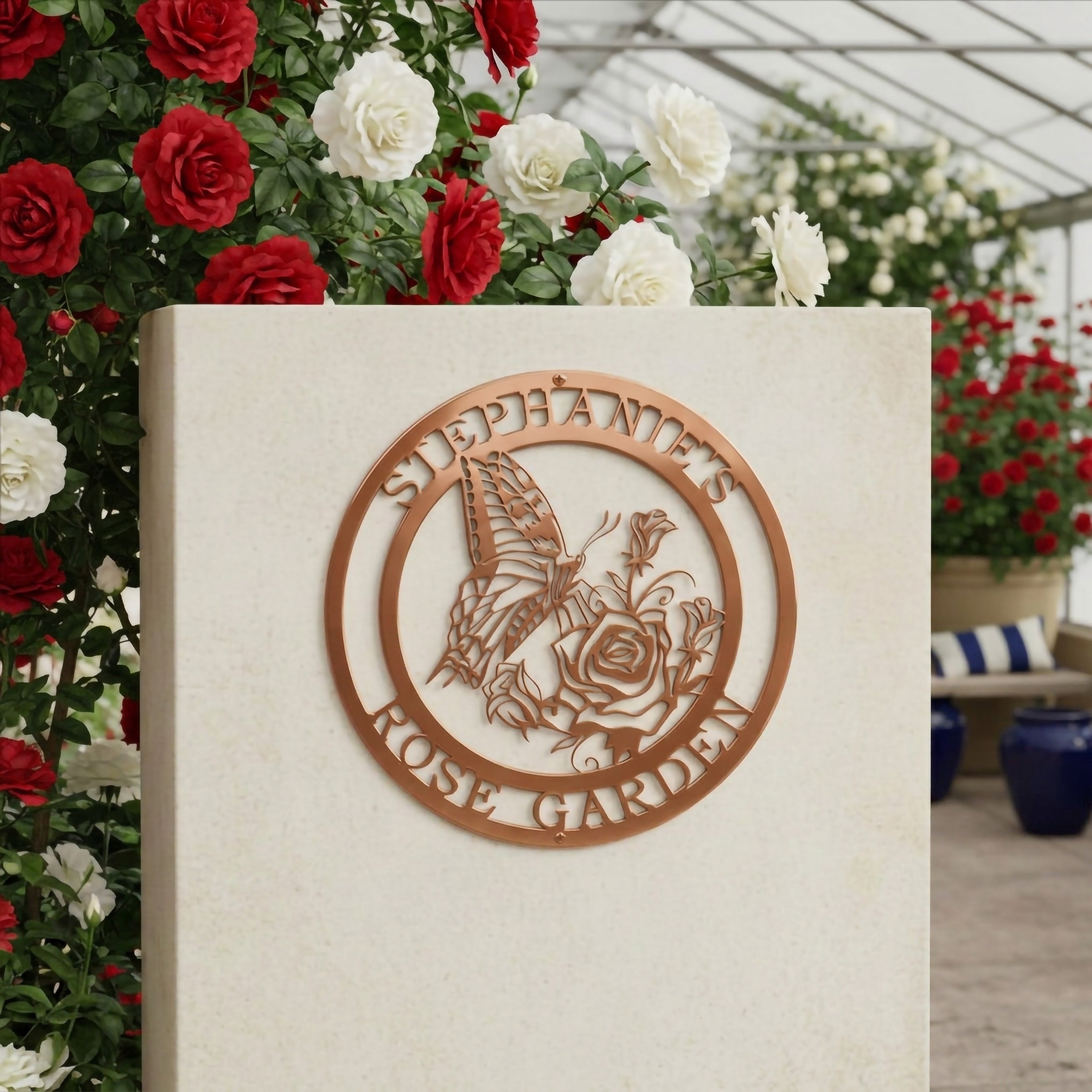 A copper-colored metallic circular sign reading "Stephanie's Rose Garden," displayed on a smooth light stone pedestal inside a greenhouse surrounded by vibrant red and white roses.