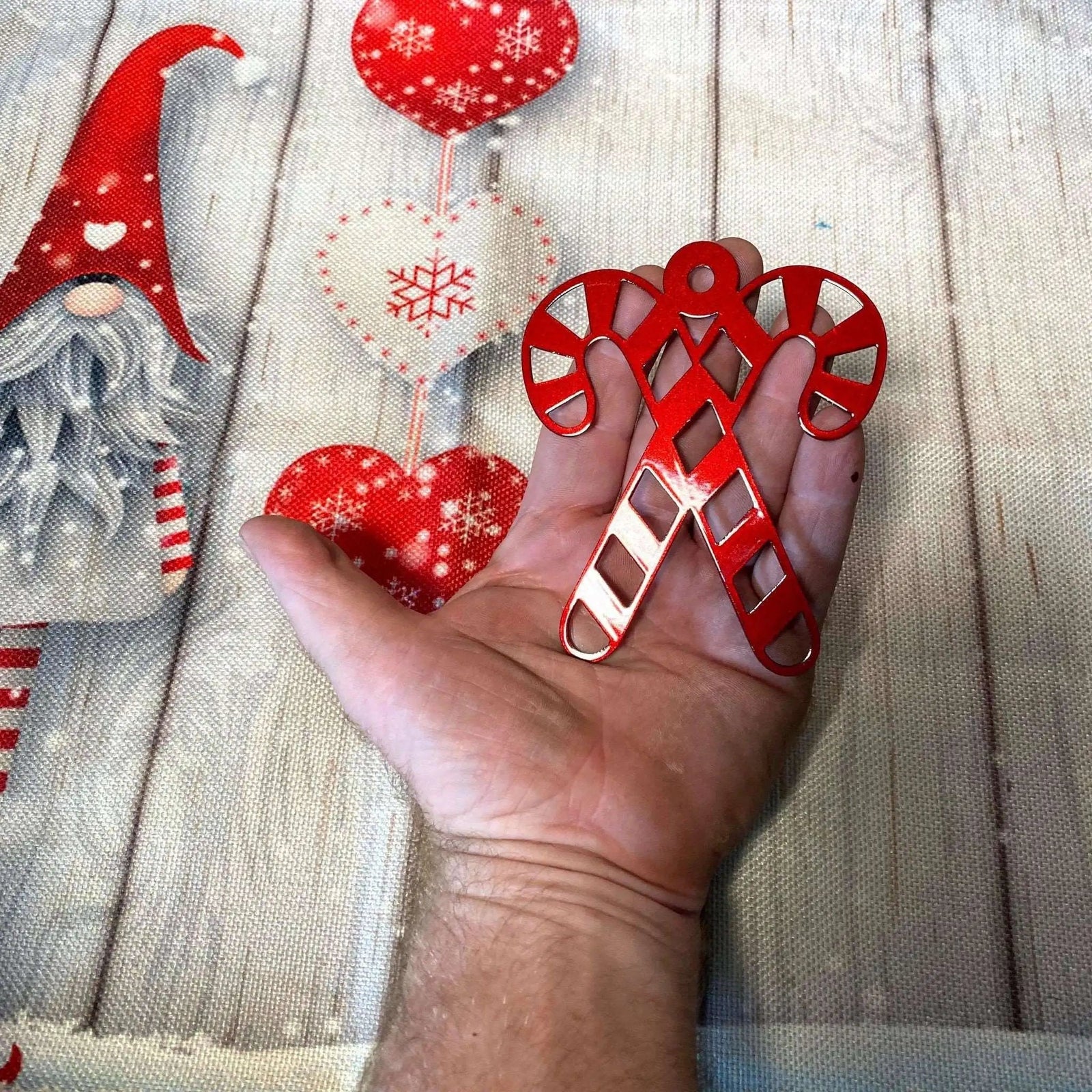 Hand holding red candy cane-shaped clip against wooden background with Christmas decorations, festive holiday accessory for hanging ornaments or seasonal décor