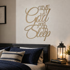 A desert tan powder-coated metal script sign reading "Give It to God and Go to Sleep" is centered on a light-colored bedroom wall above a dark wooden headboard. A dark blue quilted bed with a striped throw pillow sits next to a nightstand arranged with a Holy Bible and a lit candle lantern.