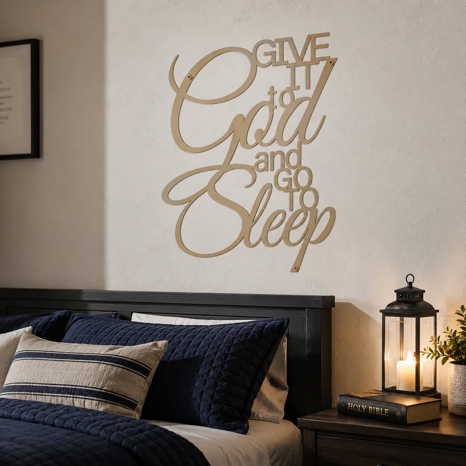A desert tan powder-coated metal script sign reading "Give It to God and Go to Sleep" is centered on a light-colored bedroom wall above a dark wooden headboard. A dark blue quilted bed with a striped throw pillow sits next to a nightstand arranged with a Holy Bible and a lit candle lantern.