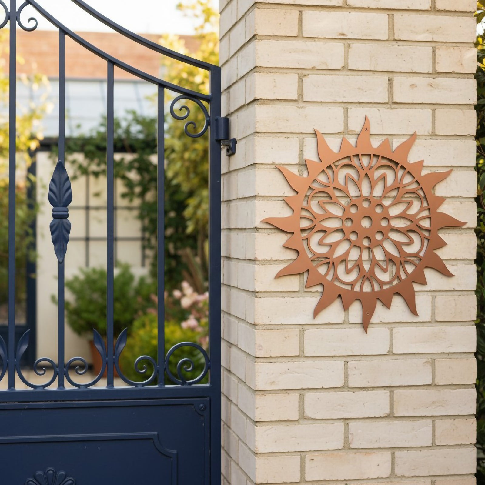  A copper-colored metallic sunburst medallion fixed to a light brick entryway pillar next to an ornate navy blue wrought-iron privacy gate.