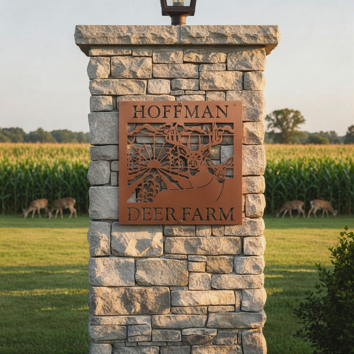 Rustic brown custom deer farm metal sign with deer and sunrise design reading “HOFFMAN DEER FARM” mounted on a stone pillar in front of a grassy field and cornfield.
