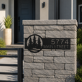 Black metal personalized address sign with forest tree cutout design mounted on gray stone gate pillar displaying 5774 Main Street