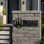 Black metal personalized address sign with forest tree cutout design mounted on gray stone gate pillar displaying 5774 Main Street