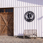Personalized cowboy and cowgirl metal farm sign mounted on white barn wall beside wooden bench outdoors