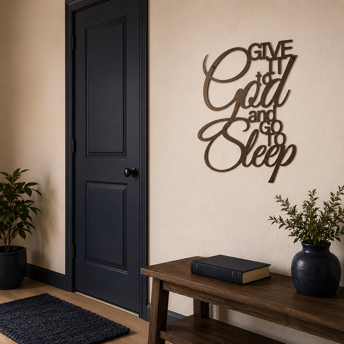 A metal wall sign with a dark oil rubbed bronze finish displaying the quote "Give It to God and Go to Sleep" is hung on a smooth beige wall. The scene features an open-concept entryway or bedroom next to a dark charcoal gray panel door and a wooden console bench with a potted plant.