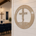 An ivory or cream-colored circular metal sign with a cut-out design of a leafy cross and the text "JOE & STEPHANIE EST 2021" is fixed to a white shiplap wall inside a church sanctuary. Rows of navy blue chairs are visible in the background leading toward an altar with a large wooden wall cross.