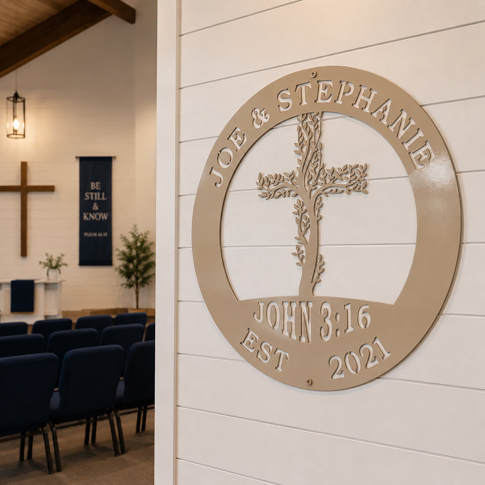 An ivory or cream-colored circular metal sign with a cut-out design of a leafy cross and the text "JOE & STEPHANIE EST 2021" is fixed to a white shiplap wall inside a church sanctuary. Rows of navy blue chairs are visible in the background leading toward an altar with a large wooden wall cross.
