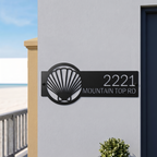 Black metal seashell address sign with house number on white wall near beach entrance