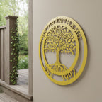A bright mustard yellow circular metal cutout sign customized with the text "THE GRANTHAMS EST. 1992" around a central tree motif. The piece is hanging on a beige exterior wall column beside a wooden porch railing wrapped in climbing green ivy.