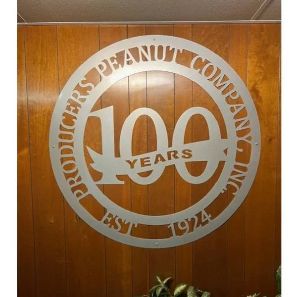 Polished silver laser-cut metal sign with “100 YEARS” at center, company name and “EST. 1924” around border, on wood wall.